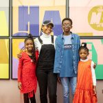 A mum and her three children stand smiling, arms around each other, in front of a large, colourful Ha Ha Hackney poster