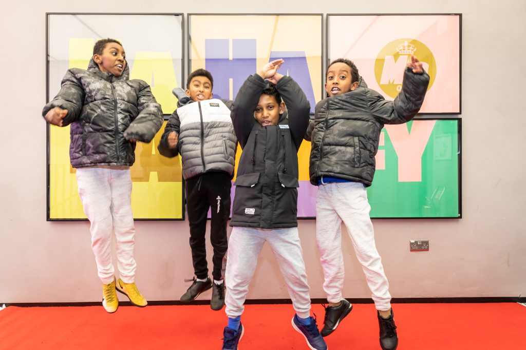 Four young teenage boys, wearing white jeans and puffer jackets jump up high in front of a large, colourful Ha Ha Hackney poster