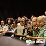 Adults smile and laugh sitting in the cinema chairs watching the young children's work