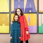 Two young girls, in bright, colourful dresses stand with their arms around each other smiling in front of a colourful Ha Ha Hackney poster