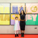 A young girl in a rainbow dress and her mum in a black dress stand smiling posed with their arms raised above their heads like ballerinas in front of a large, colourful Ha Ha Hackney poster