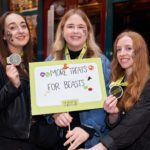 Three young woman stand smiling at the camera holding a Monster Match placard reading More Treats for Beasts.