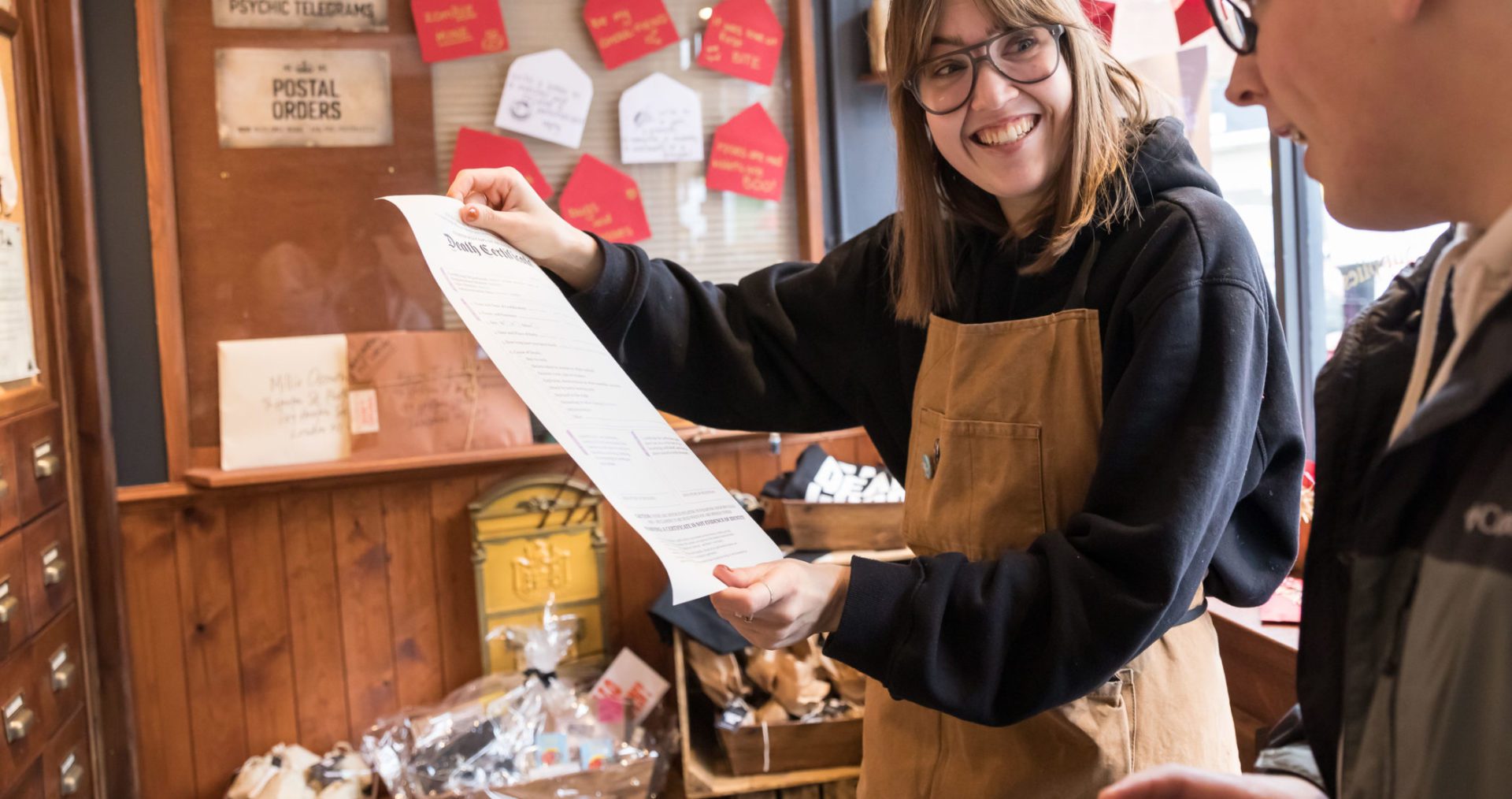 Lina is a white woman, with shoulder length brown hair and a fringe. She wearing large glasses, a shopkeepers apron and has been photographed holding a shop product. She is smiling broadly.