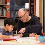 Volunteer sits with young writer and points to paper child writes in book. Book Bot in background