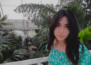 Joanna, with long dark hair, stands looking directly at the camera wearing a light green top.