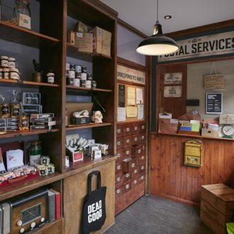 A view of the interior of the monster store. Wooden shelves along the walls stacked high with jars and bottles. A grey floor and old style hanging lights.