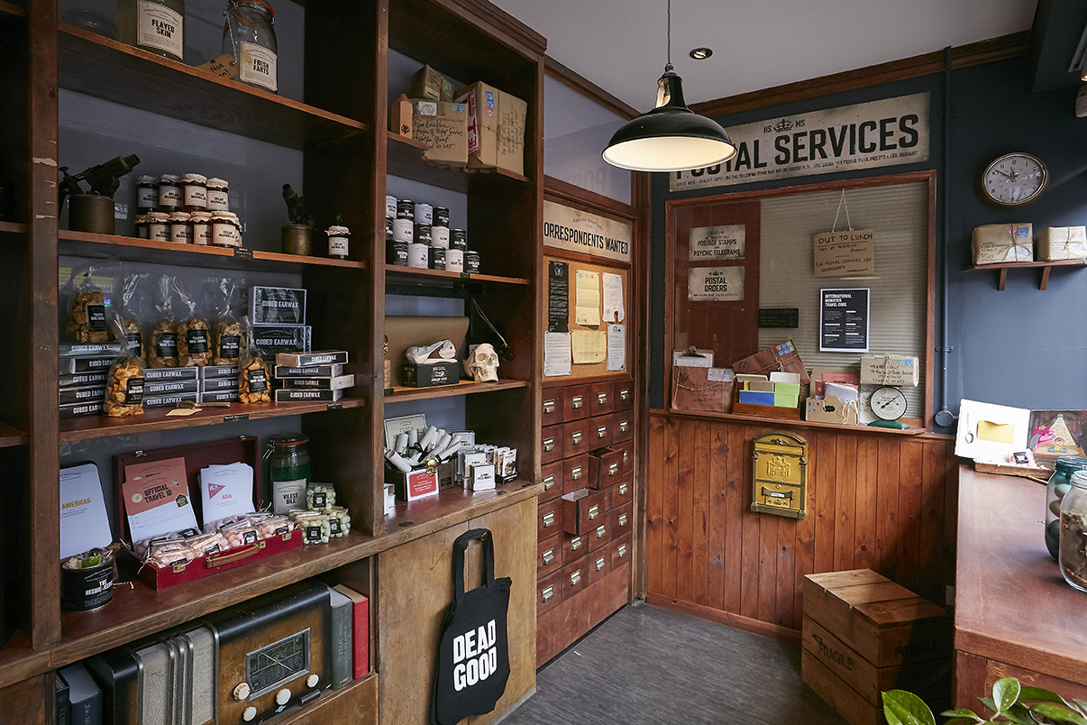 A view of the interior of the monster store. Wooden shelves along the walls stacked high with jars and bottles. A grey floor and old style hanging lights.