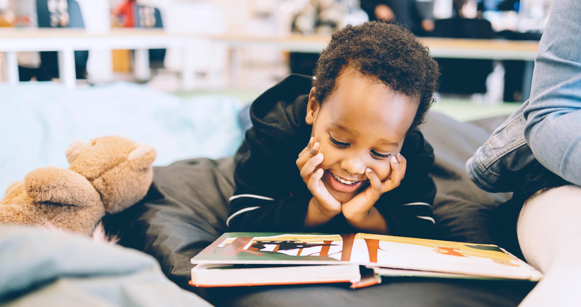 A young boy lays on his tummy, hand under his chin, smiling broadly while reading a book laid out in front of him.