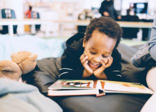 A young boy lays on his tummy, hand under his chin, smiling broadly while reading a book laid out in front of him.