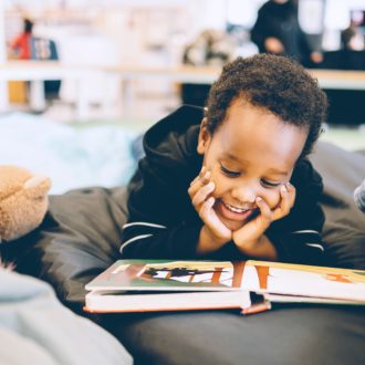 A young boy lays on his tummy, hand under his chin, smiling broadly while reading a book laid out in front of him.
