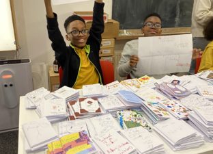 A joyful young writer in a yellow t-shirt holds their zine up above their head showing it proudly to the photographer