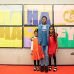 Three young people, dressed in bright clothing, stand smiling, arms around each other, in front of a large cinema poster that reads Ha Ha Hackney with the Ministry of Stories logo