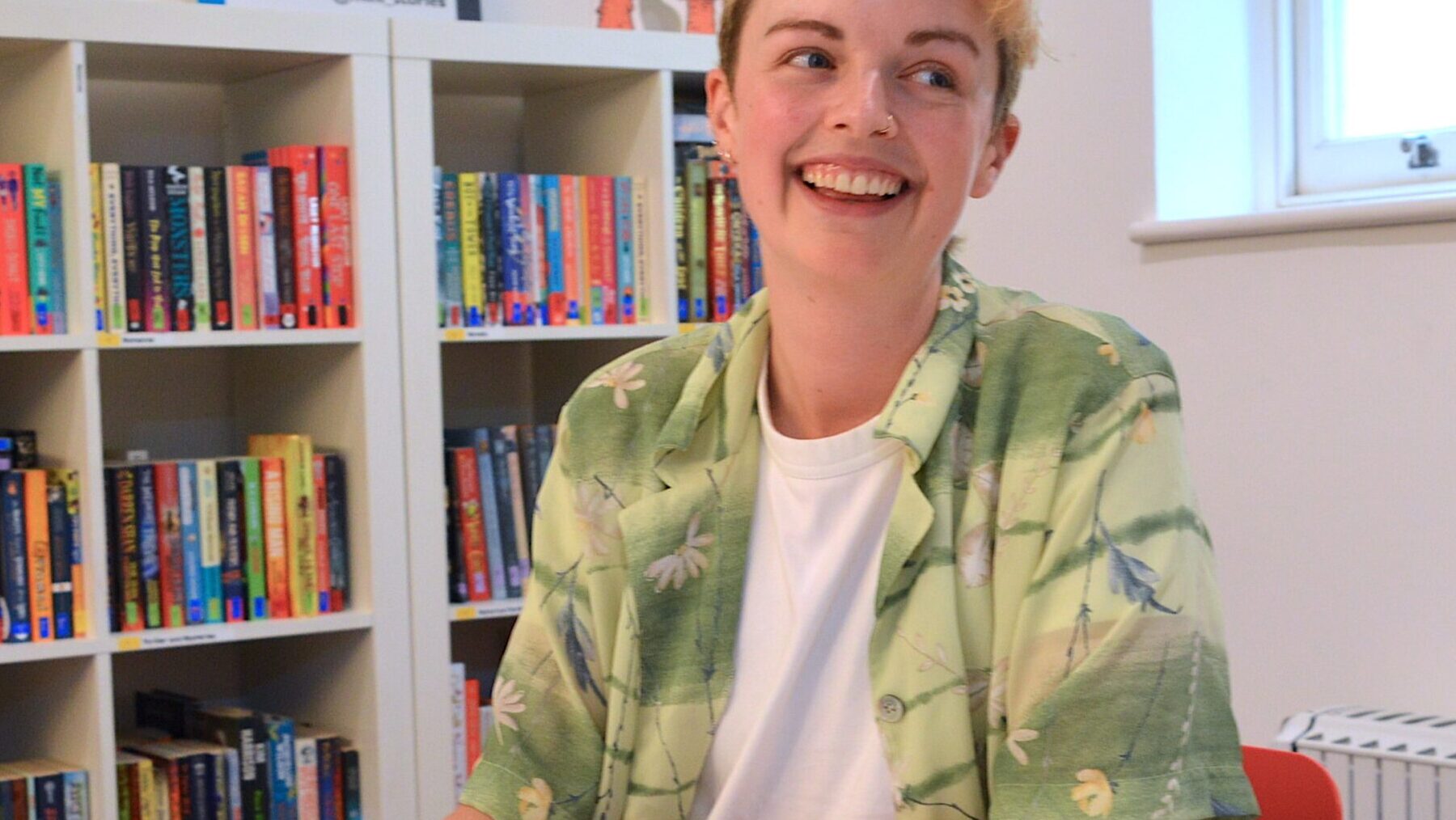 Autumn Sharkey is a white person with short bleach blonde hair. They are wearing a green short sleeved shirt over a white t-shirt and smiling looking off to the side.