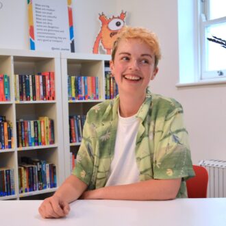 Autumn Sharkey is a white person with short bleach blonde hair. They are wearing a green short sleeved shirt over a white t-shirt and smiling looking off to the side.