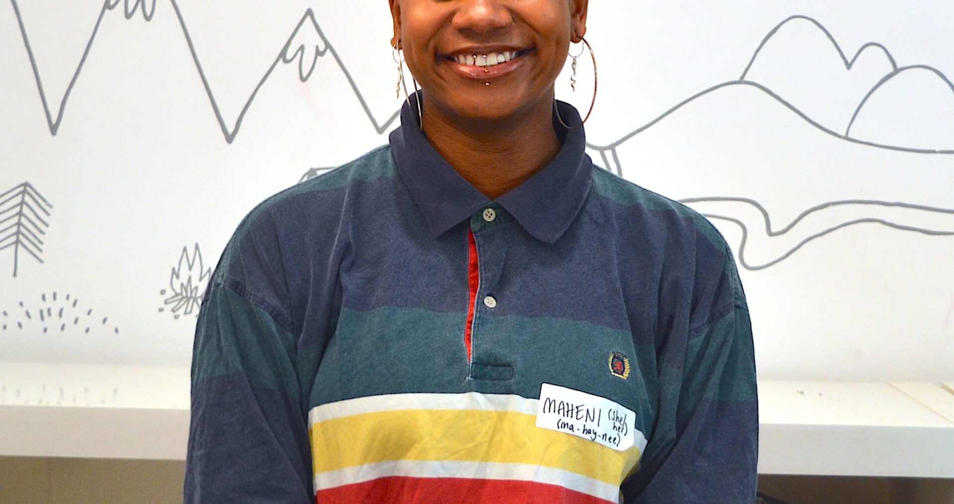 Maheni sits behind a white desk smiling broadly at the camera. She is black with a buzzcut hairstyle and is wearing two pairs of big earrings. She has a lip piercing and is wearing blue eyeshadow. Maheni is in a colourful, striped rugby shirt.