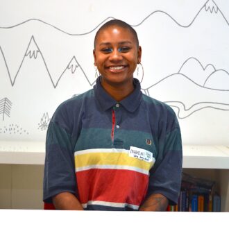 Maheni sits behind a white desk smiling broadly at the camera. She is black with a buzzcut hairstyle and is wearing two pairs of big earrings. She has a lip piercing and is wearing blue eyeshadow. Maheni is in a colourful, striped rugby shirt.