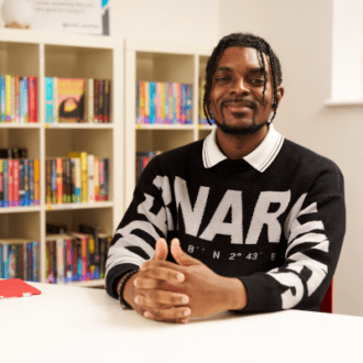 Regan - a black man with dark twists hair - sits with his hands folded in front of him. He wears a dark jumper with white writing on it and a white collar. He smiles happily at the camera.