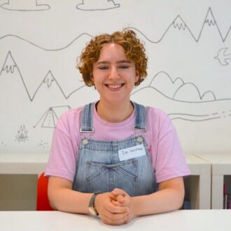 Zoe sits with their hands clasped smiling at the camera. She is a white woman with short curly hair, gold hoop earrings and a watch on her right wrist. They are wearing a lilac purple t-shirt and light blue denim dungarees.