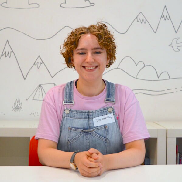 Zoe sits with their hands clasped smiling at the camera. She is a white woman with short curly hair, gold hoop earrings and a watch on her right wrist. They are wearing a lilac purple t-shirt and light blue denim dungarees.
