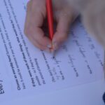 A hand holding a red pencil writes on a piece of white paper with lots of instructions on how to write about heros