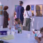 A group of children sit around a white table covered with pencils and paper while they write and talk with one another.