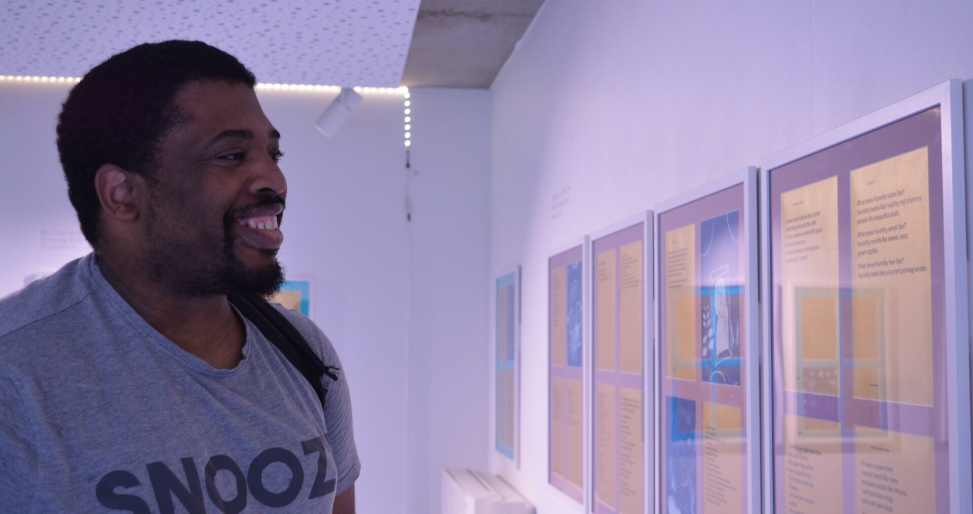 A young black man wearing a white t-shirt smiles broadly while looking at the writing displayed on the wall in front of him