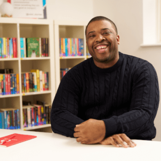 Mayo - a black man with short dark hair - sits with his hands clasped in front of him. He wears a dark navy jumper and grins widely towards the camera.