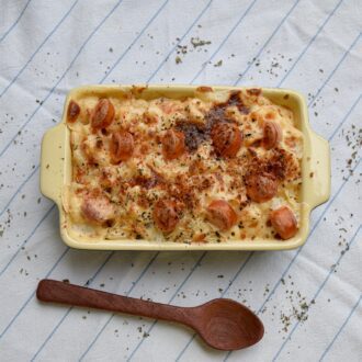 A full and tasty lasagna, bubbling with cheese and tomato, sits in a traditional, cream oven dish in the centre of a table. The table is covered in a white linen cloth with blue strips. Next to the oven dish is a brown, wooden cooking spoon and a few sprinkles of black pepper.
