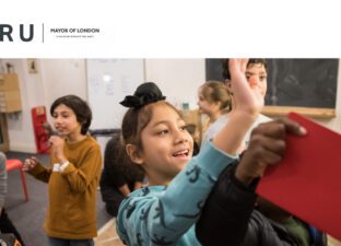 A young boy in a green jumper smiles and waves at a writing mentor out of shot. He stands in a small bring room with a big rug. The room is busy, full of children aged around 8-9 years old, talking, writing and laughing.