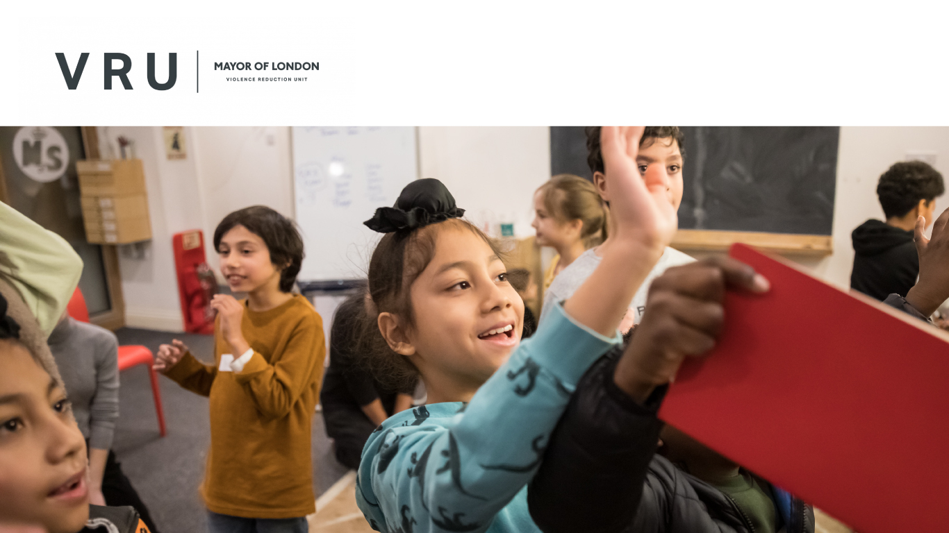 A young boy in a green jumper smiles and waves at a writing mentor out of shot. He stands in a small bring room with a big rug. The room is busy, full of children aged around 8-9 years old, talking, writing and laughing.