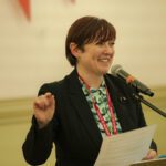 Dorothy Cotter, Ministry of Stories Education Programme Manager with short red hair, black blazer and a bright green patterned shirt, smiles out and addresses the crowd from the lectern.