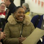 A member of the audience - a young woman, with dark hair, large gold earrings and a pale puffer coat with fur hood, smiles widely at a young person off camera.