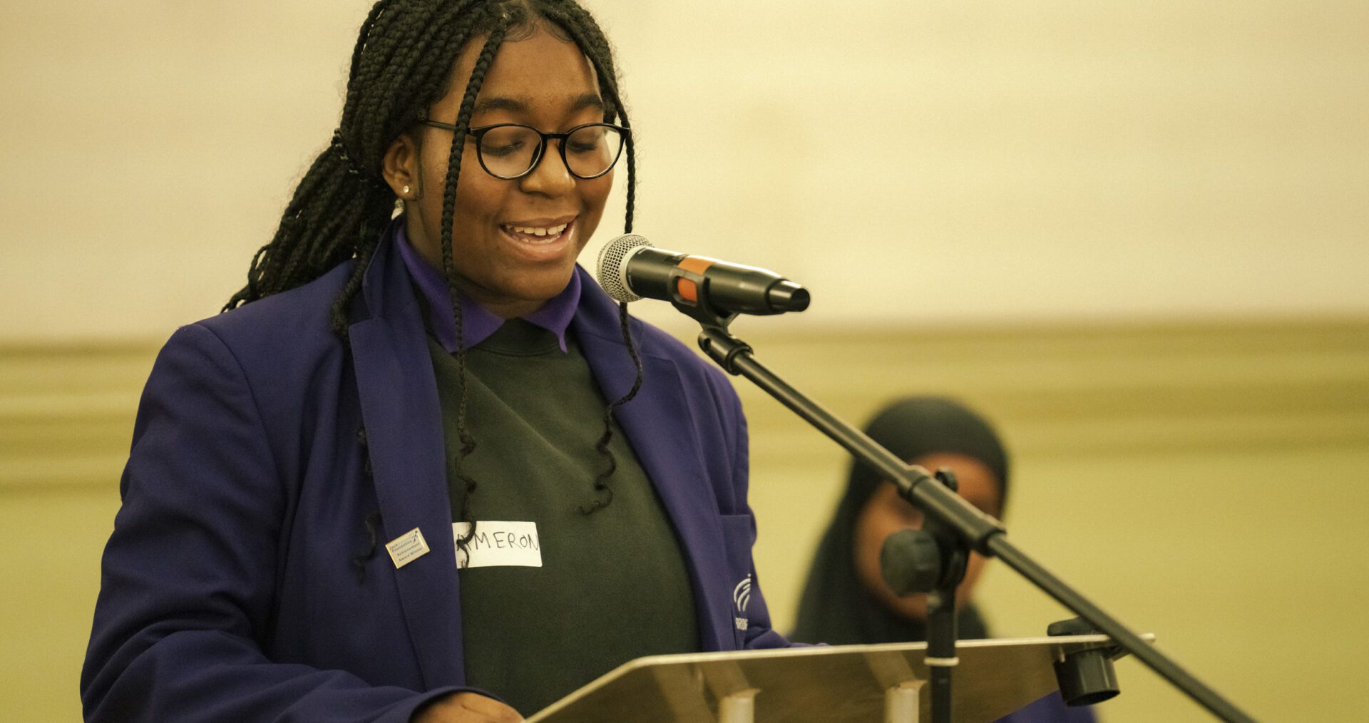 A teen girl with braids, dark glasses and a purple blazer smiles as she stands at the lectern and delivers her speech to the crowd