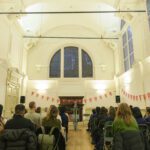 A large group of people are seated in a large white room, with heigh ceilings and a large, ornate window on the back wall. Red Ministry of Stories bunting zig zags across the room.