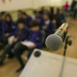 Close up view of a microphone standing on top of a lectern. There is a large room in front of the stand and out of focus, sits many students dressed in purple blazers