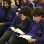 Two teenage boys, with dark curly hair and bright purple hair sit together, heads together looking at white sheets of paper in their hands, practicing their speeches