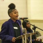 A teen girl with dark hair in a large afro bun on top of her head, wearing a purple blazer, speaks animatedly to the crowd ahead of the lectern she stands in front of