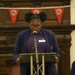 A teen girl, wearing dark glasses, a purple blazer and dark hair in large space buns, stands in the lectern delivering her speech.