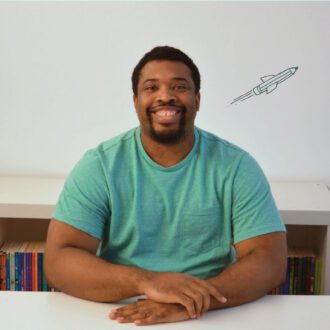 Mayo, a black man with dark hair and a trimmed beard, sits smiling at the camera his hand clasped in front of him. He wears a bright, teal t-shirt and has a small illustrated rocket blasting off on the wall behind him.