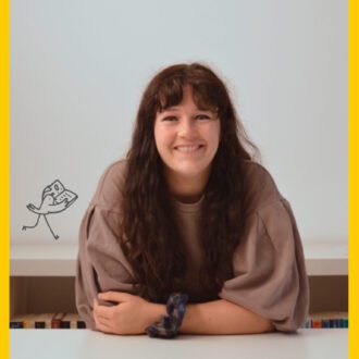 Jenny, a white woman with long brunette hair and a fringe, sits smiling a the camera, arms folded in front of her. She wears a light brown sweater with large sleeves and has a hair scrunchie around her wrist.