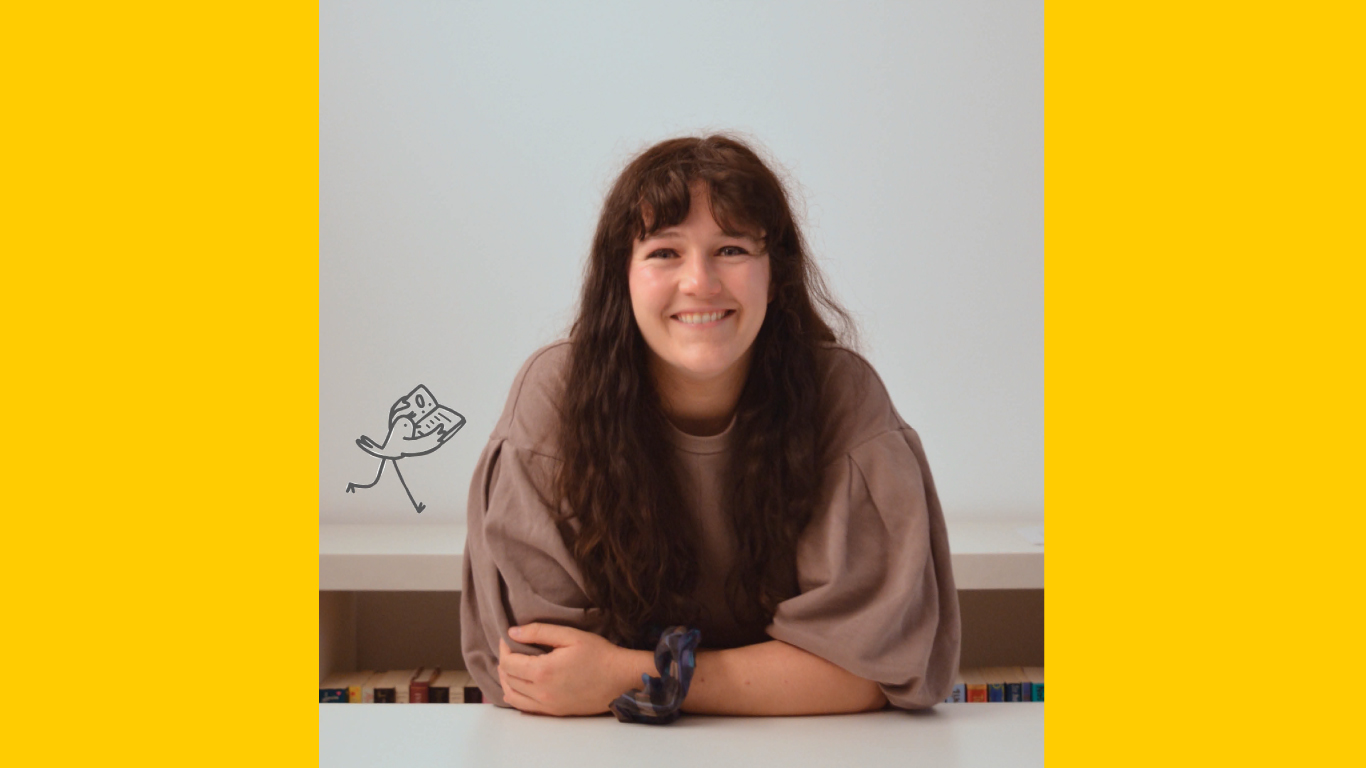 Jenny, a white woman with long brunette hair and a fringe, sits smiling a the camera, arms folded in front of her. She wears a light brown sweater with large sleeves and has a hair scrunchie around her wrist.