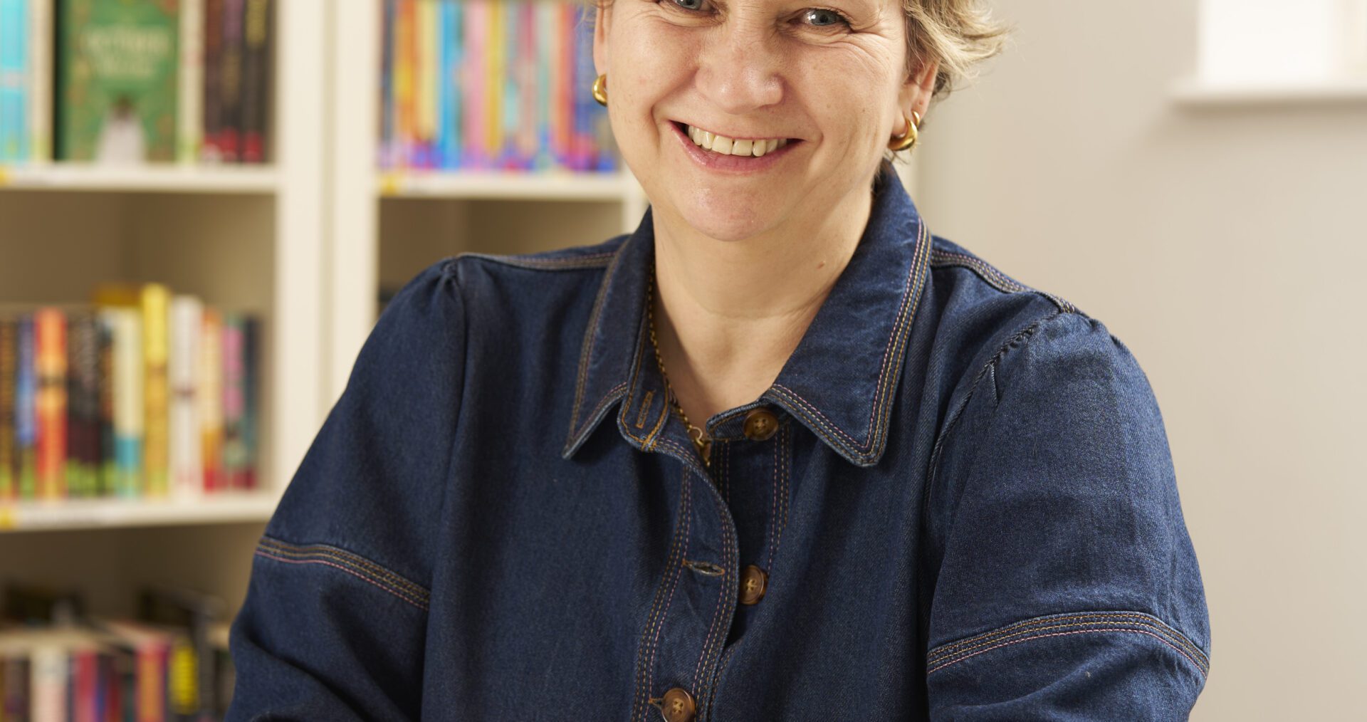 Jane - a white woman with short, blonde hair - its with her hands folded in front of her. She wears a dark denim shirt and gold earrings and smiles happily at the camera.