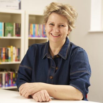 Jane - a white woman with short, blonde hair - its with her hands folded in front of her. She wears a dark denim shirt and gold earrings and smiles happily at the camera.