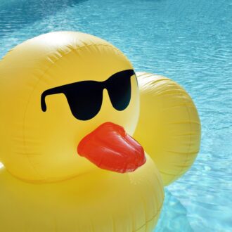 A large inflatable, yellow duck wearing sunglasses floats alone on a large blue swimming pool