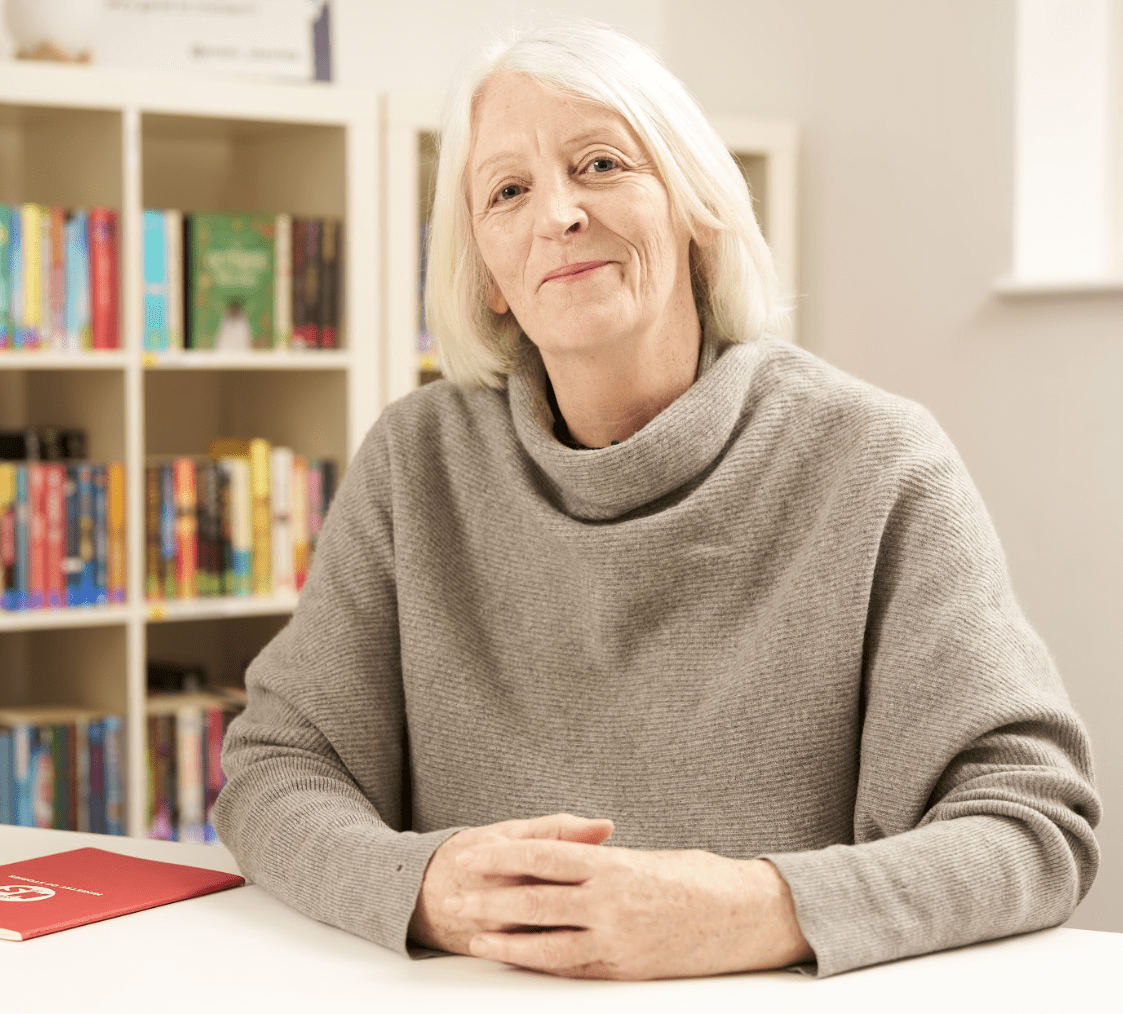 Cath Greenwood - a white woman with blonde hair, shoulder length hair - site hands clasped in front of her. She wears a taupe, cowl neck jumper and smiles at the camera.