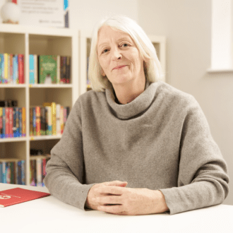 Cath Greenwood - a white woman with blonde hair, shoulder length hair - site hands clasped in front of her. She wears a taupe, cowl neck jumper and smiles at the camera.