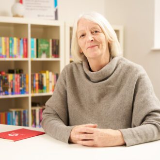 Cath Greenwood - a white woman with blonde hair, shoulder length hair - site hands clasped in front of her. She wears a taupe, cowl neck jumper and smiles at the camera.