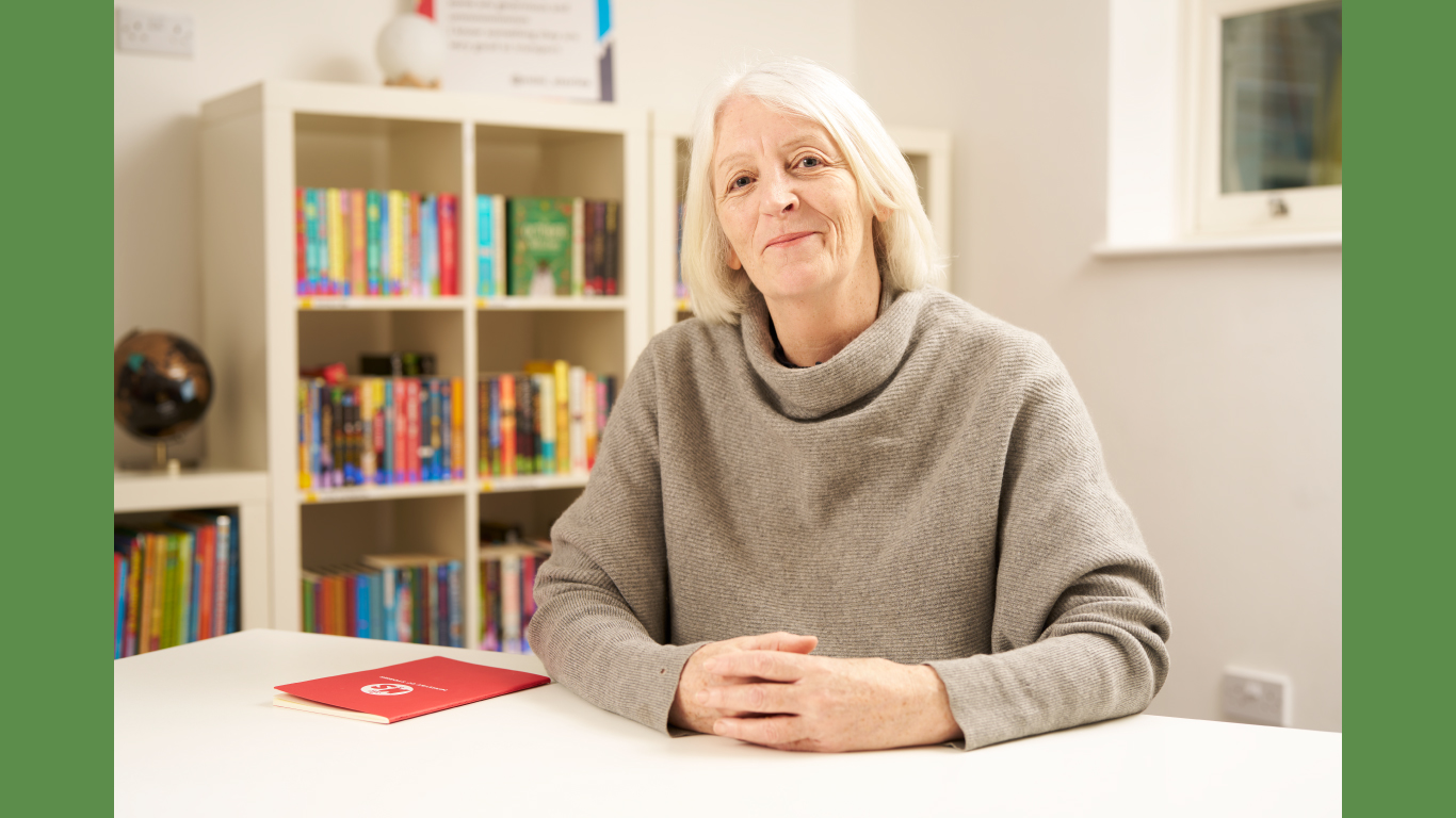 Cath Greenwood - a white woman with blonde hair, shoulder length hair - site hands clasped in front of her. She wears a taupe, cowl neck jumper and smiles at the camera.