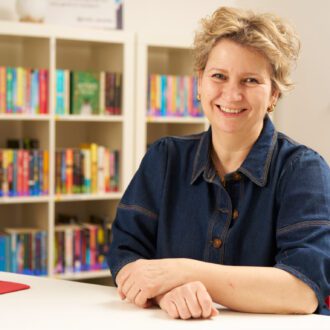 Jane - a white woman with short, blonde hair - its with her hands folded in front of her. She wears a dark denim shirt and gold earrings and smiles happily at the camera.