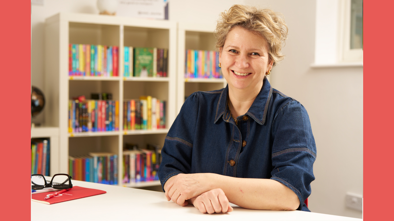 Jane - a white woman with short, blonde hair - its with her hands folded in front of her. She wears a dark denim shirt and gold earrings and smiles happily at the camera.